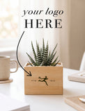 A small potted succulent in a light wood rectangular planter sits on a white tabletop in a bright indoor setting. The planter is engraved with a soccer player silhouette and the text 'FTD 1910,' with overlaid text reading 'your logo HERE' and an arrow pointing to the engraving area. - Image 