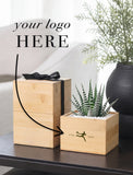 Two personalized bamboo gift boxes on a dark table, one stacked with a black ribbon and one holding a small potted succulent. The image includes a placeholder text 'your logo HERE' and a black arrow pointing to the box design. - Image 
