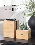 Two light natural wood gift boxes on a dark tabletop, with one box tied by a black ribbon and the other a square planter holding a small striped succulent. Large text reads 'your logo HERE' with an arrow pointing to the front box, which also has a small green logo and 'FTD 1910' printed on it. - Image 