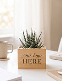 A small potted succulent in a light wood square planter box printed with the placeholder text 'your logo HERE', sitting on a white desk in a bright, softly blurred indoor setting. - Image 