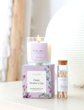 Burning white candle in glass jar with "Petal Blush" label, on a purple "Happy Mother's Day" box, next to a glass vial of white matches. - Image 
