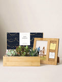 A wooden photo frame with a cork board and sticky notes stands beside a bamboo planter box containing succulents, with a navy holiday card in the background. The cards and frame show the brand name 'L. - Image 