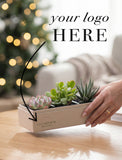 A hand holds a beige rectangular planter box containing small succulent plants on a wooden table, with a blurred holiday tree and sofa in the background. The planter has the text 'CLEARVIEW REAL ESTATE' printed on the front, and large overlaid text reads 'your logo HERE' with a black arrow pointing to it. - Image 