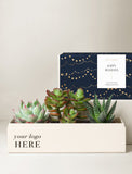 A holiday succulent gift set featuring a cream-colored wooden planter box with printed placeholder text, filled with three green succulent plants, with its navy blue and gold gift box behind it on a light beige background. - Image 