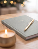 A green hardcover notebook with the word 'NOTES' in gold foil, paired with a gold pen, resting on a wooden table next to a lit candle in a gold holder. The background is softly blurred with warm bokeh lights. - Image 