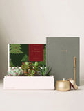 A curated gift set on a light beige surface against a neutral background, featuring a white box labeled 'LULA'S GARDEN' with succulents, a dark green notebook with gold lettering reading 'NOTES', a gold pen, a gold candle, and a Christmas-themed card. - Image 