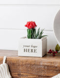 A square white planter box with the text 'your logo HERE' printed in black, styled on a rustic wooden surface with a small cactus and succulent inside. The background is a clean white paneled wall. - Image 