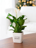 A potted peace lily plant with green leaves and white flowers in a square white planter printed with 'your logo HERE', placed on a wooden table with a blurred living room background and warm bokeh lights. - Image 