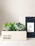 A square cream-colored succulent planter box filled with green and pinkish succulents, shown in front of its matching branded box lid. The planter reads 'your logo HERE' and the lid features a navy blue design with gold dots and a white card that says 'HAPPY HOLIDAYS'. - Image 