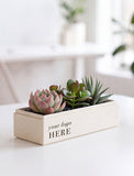 A rectangular cream-colored wooden planter box with the text 'your logo HERE' printed on the front, filled with various green and pink succulent plants, set on a white table in a bright, blurred indoor environment. - Image 