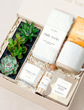 Welcome home gift box featuring three succulents, white fluffy socks, a speckled ceramic mug, tea, and a vanilla candle. - Image 
