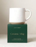 Speckled white ceramic mug with a natural base sitting on a dark green Lula's Garden gift box. - Image 