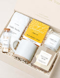 Welcome Home gift box featuring white fluffy socks, yellow herbal tea, a speckled ceramic mug, matches, and a scented candle. - Image 
