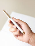 Hand holding a sleek white Lula's Garden pen with elegant gold accents and a matching clip against a soft white background. - Image 