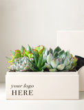 A square cream-colored gift box with printed placeholder text 'your logo HERE' on the front, filled with a small arrangement of green succulents and cacti. A matching cream box is partially visible behind the arrangement on a light neutral background. - Image 