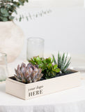 A rectangular cream-colored wooden planter box with the text 'your logo HERE' printed on the front, filled with green and purple succulent plants. The planter is styled on a white tablecloth with a blurred glass candle holder and a textured vase in the background. - Image 