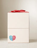 Cream-colored gift box with a red ribbon bow and a patriotic heart decal featuring stars and stripes. - Image 